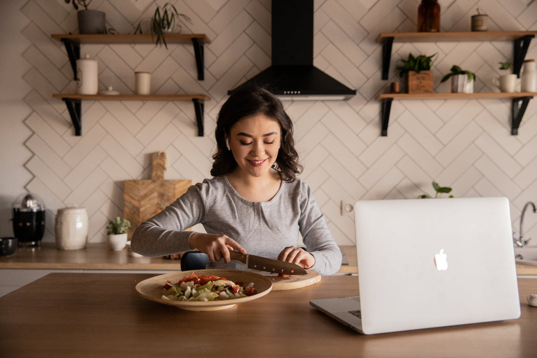 Woman cutting vegetables in her kitchen with an open laptop 